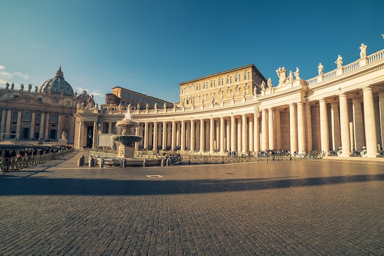 Luftfoto af Petersplads i Vatikanet, Rom, med den imponerende barokarkitektur, runde kolonnade og den majestætiske Peterskirke under klar blå himmel
