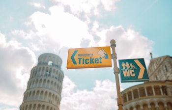 Gult vejskilt med pil der viser vej til billetkontoret i Pisas gader, nyttigt for turister der besøger det skæve tårn i Italien