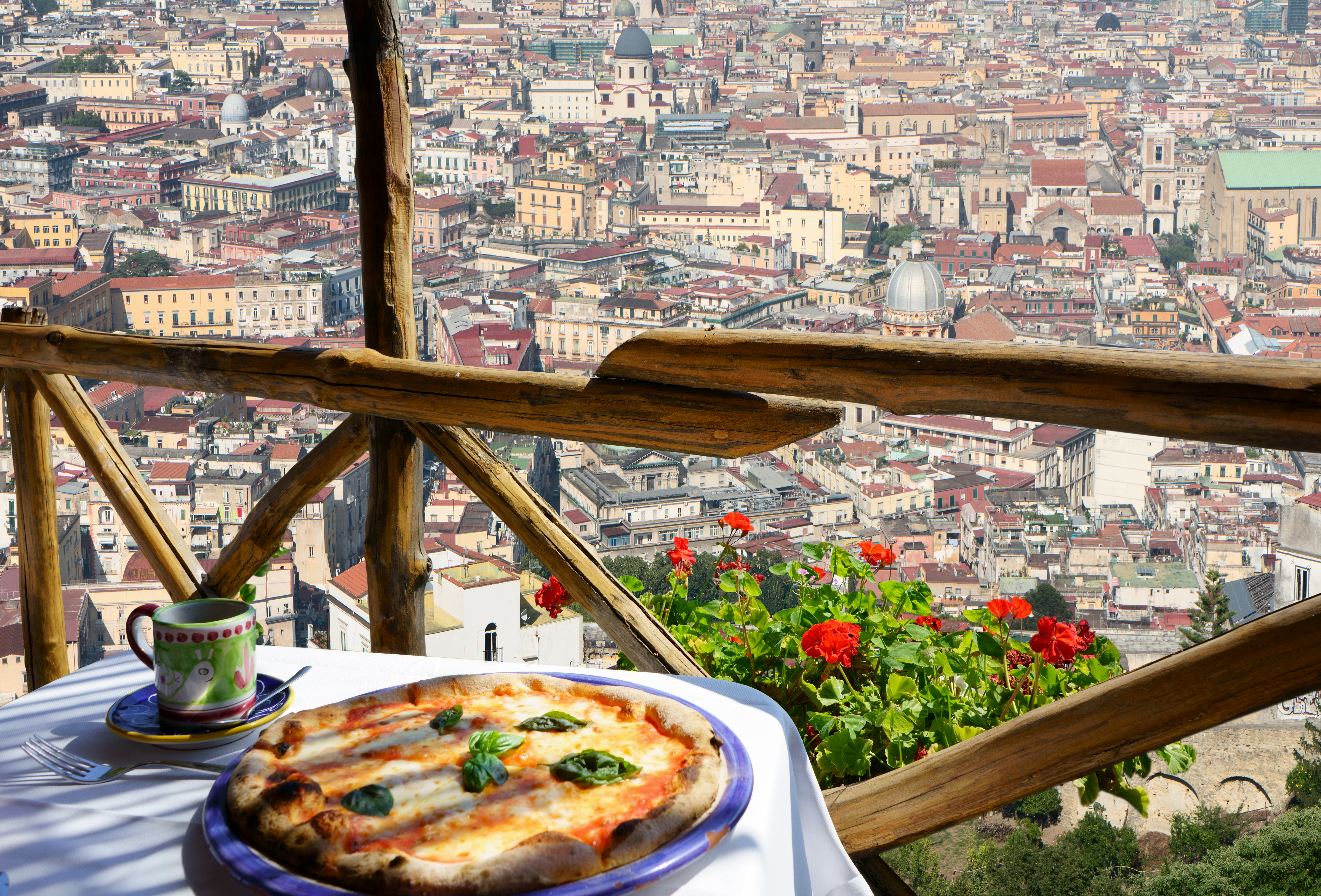 Autentisk italiensk pizza Margherita serveret på smuk terrasse med panoramaudsigt over Napoli by og røde geranier