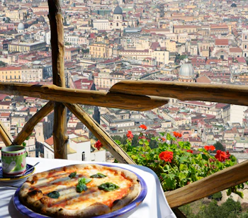 Autentisk italiensk pizza Margherita serveret på smuk terrasse med panoramaudsigt over Napoli by og røde geranier
