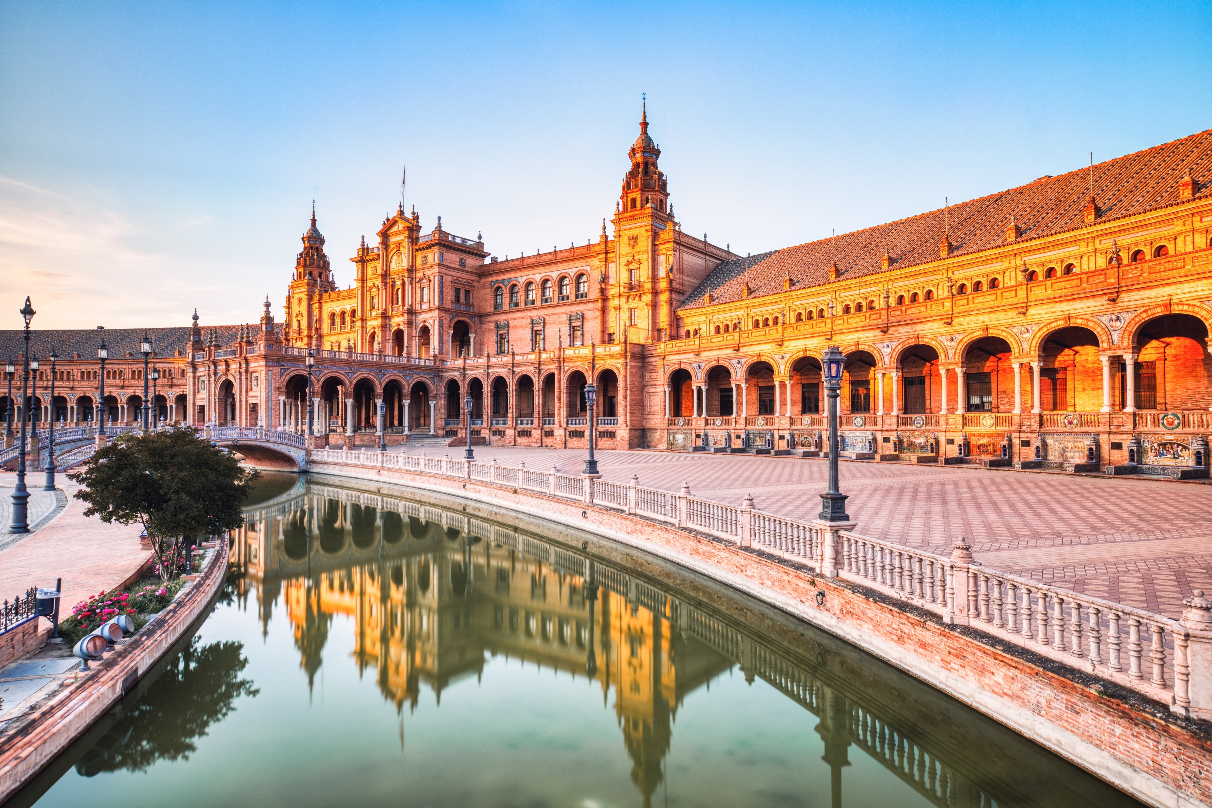Betagende udsigt over Plaza de España i Sevilla ved solnedgang med den ikoniske halvmåneformede bygning, dekorative broer og kanal, der reflekterer det gyldne aftenlys i Andalusien, Spanien