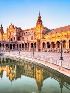 Betagende udsigt over Plaza de España i Sevilla ved solnedgang med den ikoniske halvmåneformede bygning, dekorative broer og kanal, der reflekterer det gyldne aftenlys i Andalusien, Spanien