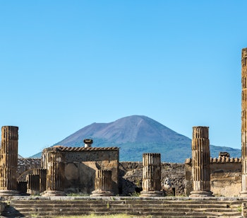 Fascinerende ruiner af den antikke romerske by Pompeji med velbevarede stensøjler og bygninger, der blev ødelagt af Vesuvs udbrud i år 79, med vulkanen majestætisk i baggrunden under en klar blå himmel