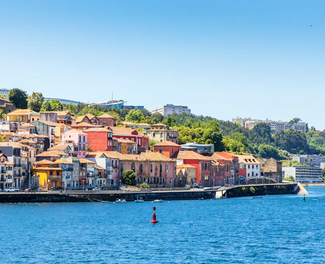 Farverige historiske bygninger langs Douro-flodens bred i Vila Nova de Gaia, Porto, Portugal, med terrakottafarvet tagsten og grønne bakkeskråninger under en klar blå himmel