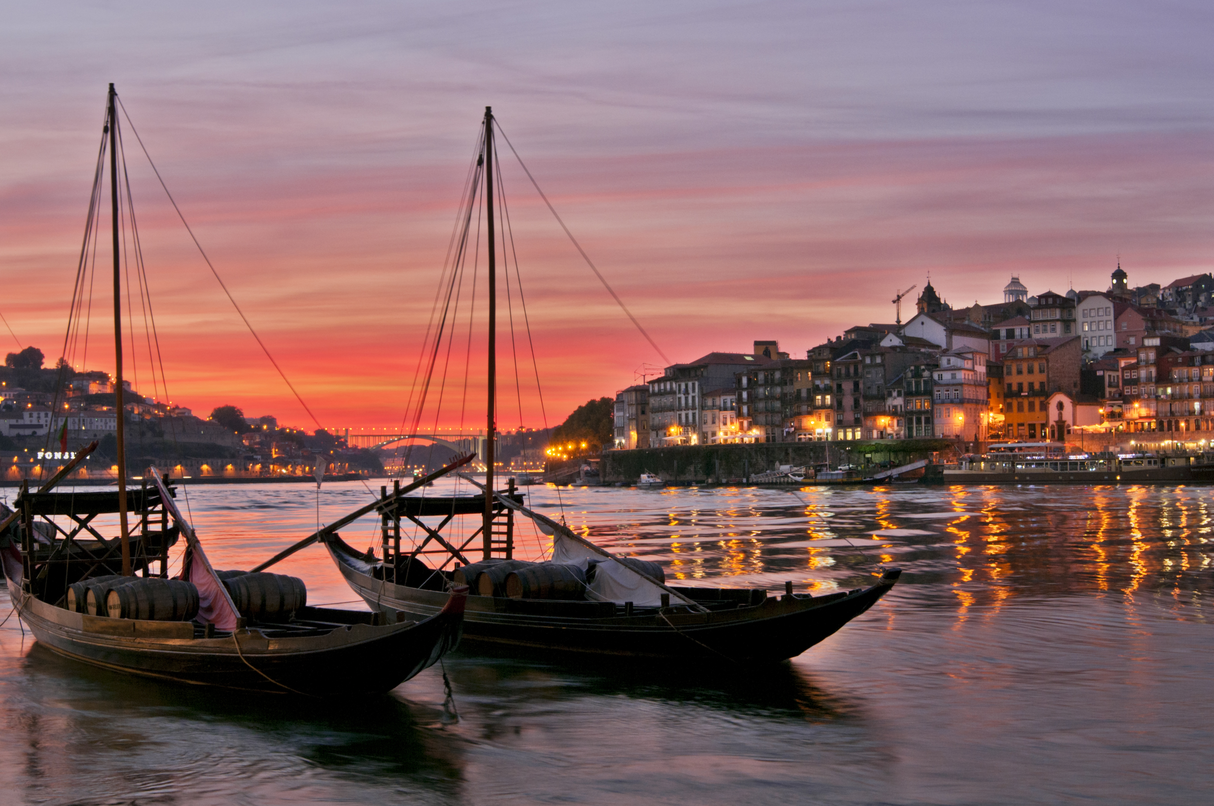 Traditionelle rabelo-både lastet med portvinsfade lægger til ved Douro-flodens bred i Porto, Portugal, mens den dramatiske røde solnedgangshimmel kaster et magisk lys over byens historiske skyline