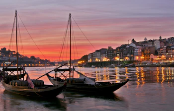 Traditionelle rabelo-både lastet med portvinsfade lægger til ved Douro-flodens bred i Porto, Portugal, mens den dramatiske røde solnedgangshimmel kaster et magisk lys over byens historiske skyline