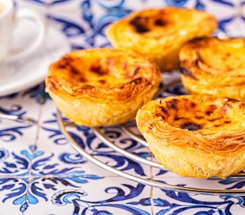 Traditionel portugisisk pastel de nata æggetærte med sprød skorpe og cremet fyld, serveret mod blå azulejo-fliser i Lissabon