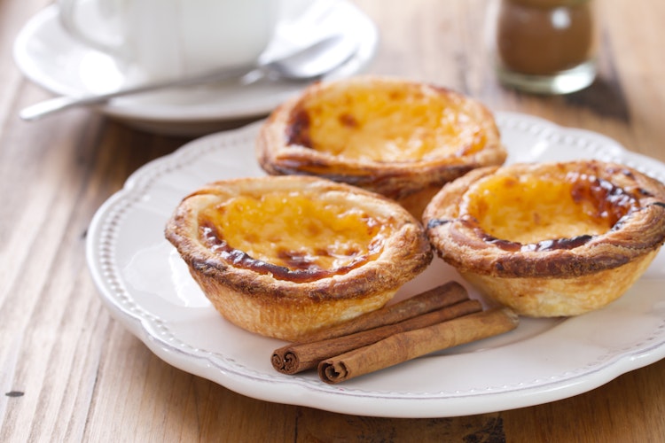 Traditionelle portugisiske Pastéis de Nata med sprød butterdej og cremet vaniljecustard, serveret med kanelstænger - en berømt dessert fra Lissabon