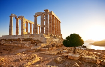 Det antikke Poseidon tempel på Kap Sounion i Grækenland badet i gyldent solnedgangslys, med majestætiske doriske søjler der troner over Det Ægæiske Hav fra sin dramatiske placering på klippetoppen.