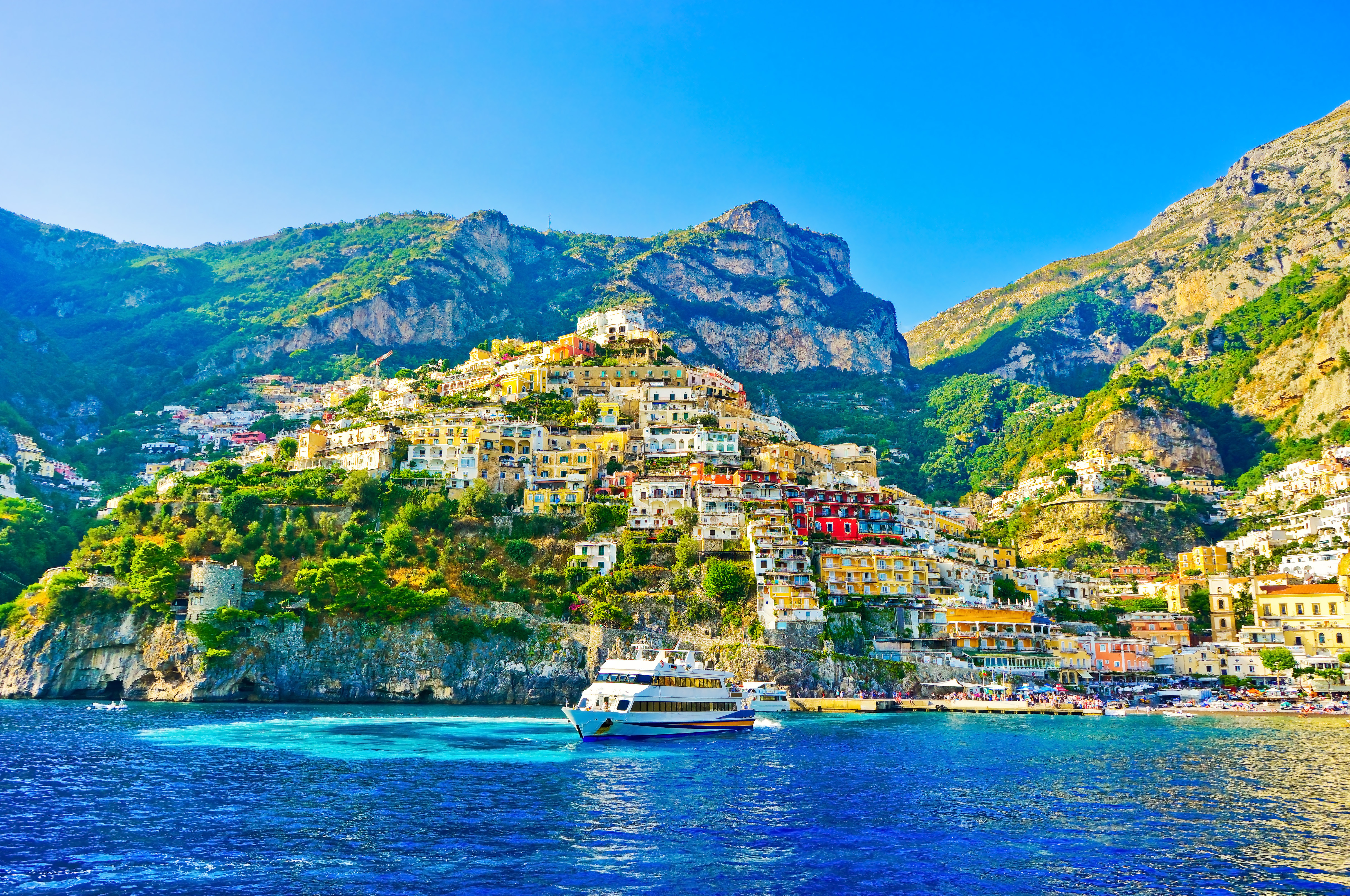 Betagende panoramaudsigt over Positano landsby paa Amalfikysten i Italien, med farverige bygninger der kaskader ned ad bjergskraaningen til en strand hvor turister slapper af ved det turkisblaae Middelhav