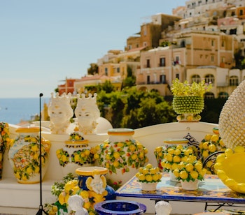 Farvestrålende traditionel napolitansk keramik i forgrunden med panoramaudsigt over Positanos ikoniske pastellfarvede bygninger, der kaskader ned ad klipperne mod Middelhavet på Amalfikysten