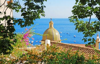 Gylden kirkekuppel i Positano med farverige blomster og udsigt over det blå Middelhav ved Amalfikysten i Italien