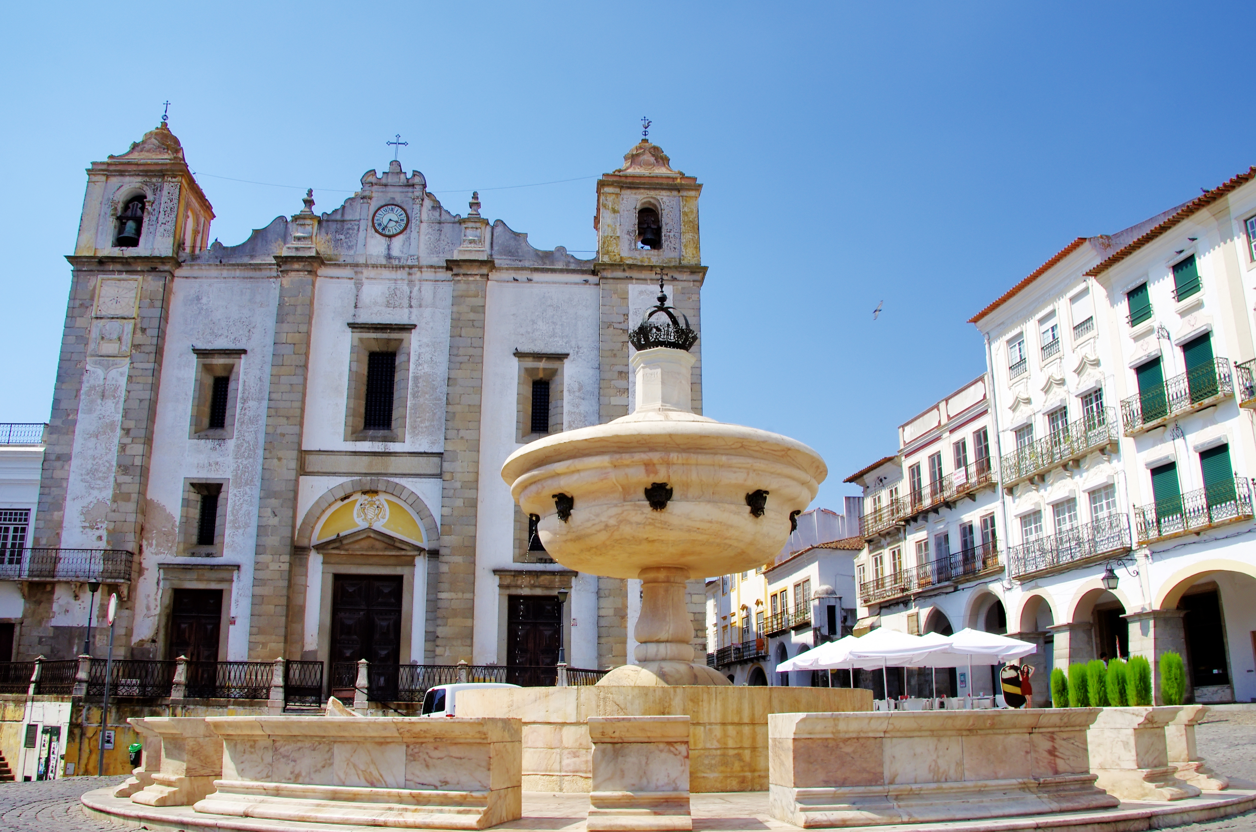 Det historiske Praça do Giraldo torv i Évora med traditionel portugisisk arkitektur, fontæne og buer under blå sommerhimmel