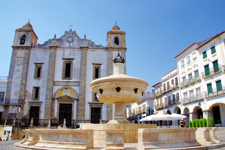 Det historiske Praça do Giraldo torv i Évora med traditionel portugisisk arkitektur, fontæne og buer under blå sommerhimmel