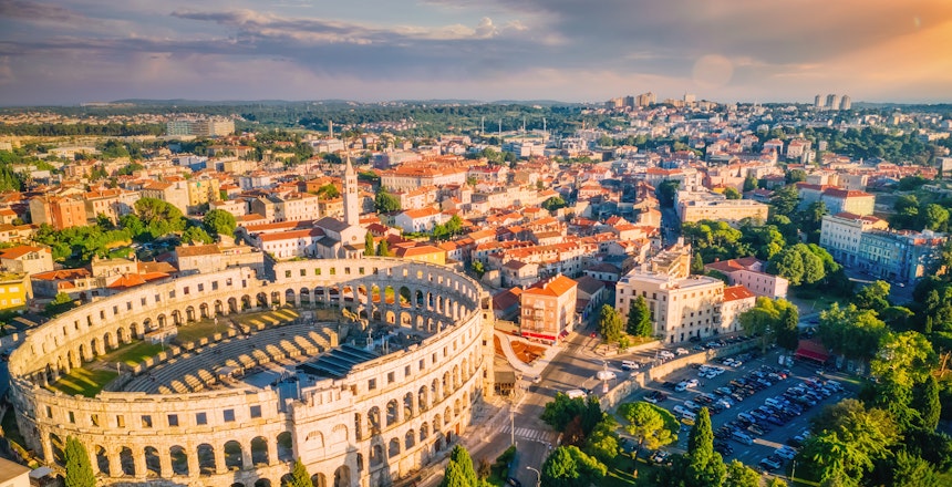 Betagende luftfoto af det antikke romerske amfiteater i Pula, Kroatien med byens panorama badet i gyldent solnedgangslys