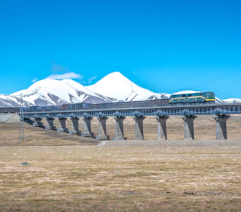 Passagertog på Qinghai-Tibet jernbanen krydser bro med snedækte Kunlun bjerge