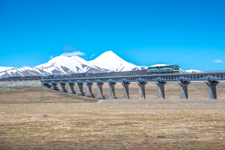 Passagertog på Qinghai-Tibet jernbanen krydser bro med snedækte Kunlun bjerge