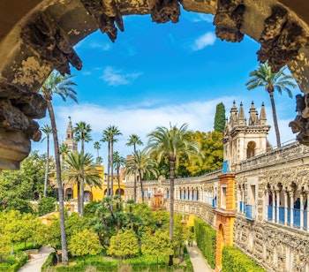 Betagende udsigt gennem en kunstfærdig bue ud mod de frodige haver og den mauriske arkitektur i Real Alcazar, Sevillas kongelige palads under blå himmel