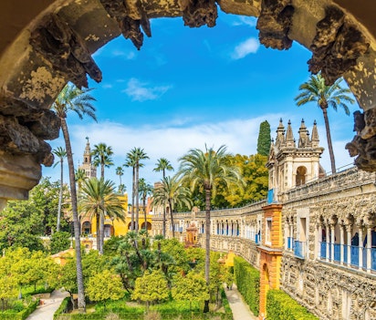 Betagende udsigt gennem en kunstfærdig bue ud mod de frodige haver og den mauriske arkitektur i Real Alcazar, Sevillas kongelige palads under blå himmel