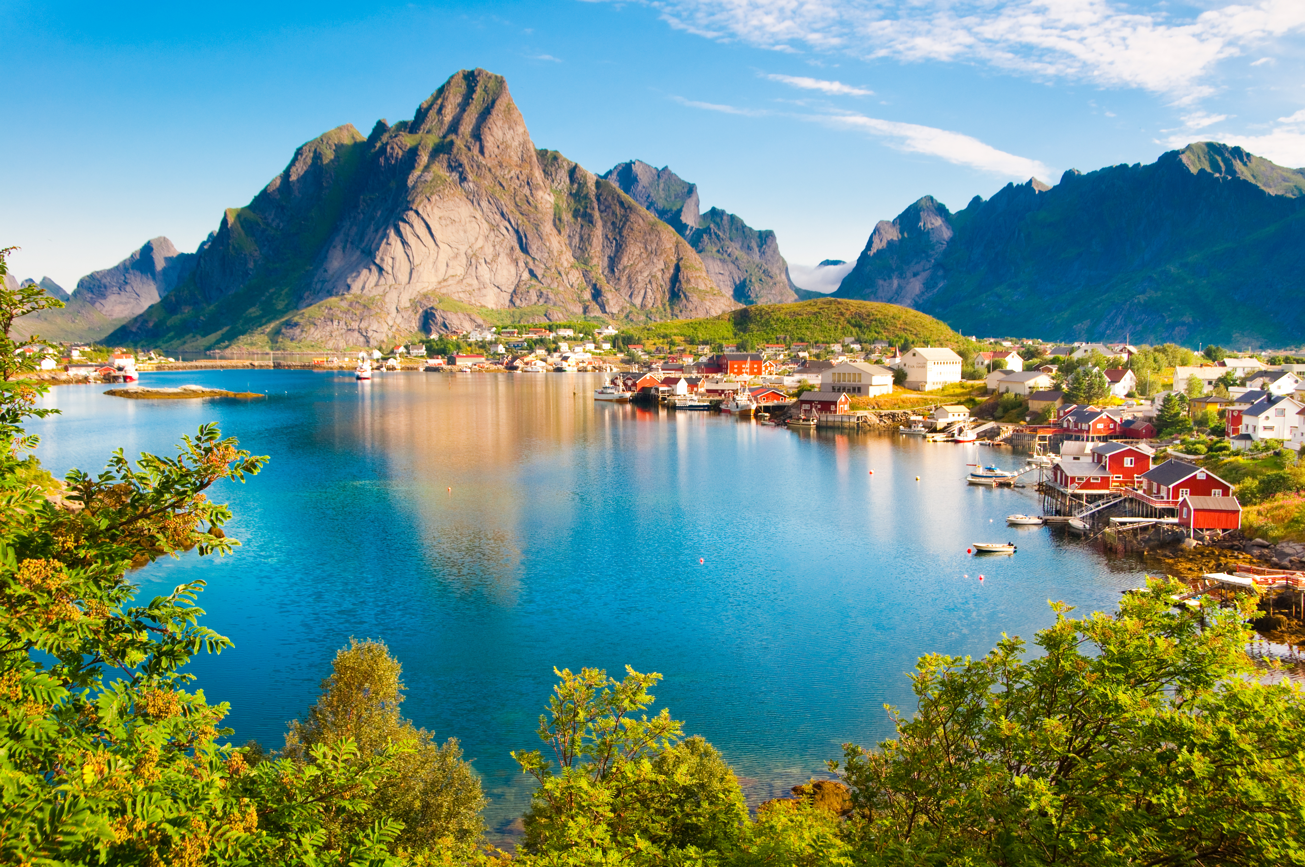 Den idylliske fiskerby Reine på Lofoten med traditionelle røde rorbuer langs vandkanten, omgivet af majestætiske bjerge og turkisblåt fjordvand i Nordnorge