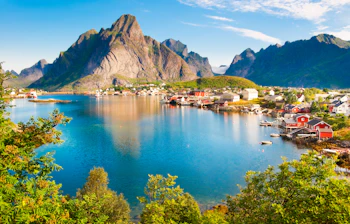 Den idylliske fiskerby Reine på Lofoten med traditionelle røde rorbuer langs vandkanten, omgivet af majestætiske bjerge og turkisblåt fjordvand i Nordnorge