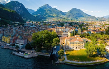 Luftfoto af Riva del Garda ved Gardasøens nordlige bred med farverige historiske bygninger, marina med både og majestætiske bjerge i baggrunden