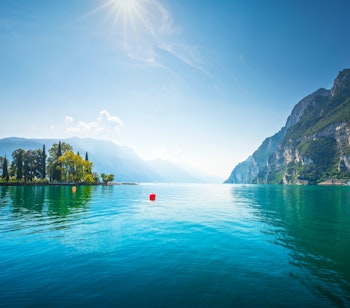 Idyllisk udsigt over Riva del Garda med frodige haver og træer langs Gardasøens bred med bjerge i baggrunden i Trentino-regionen