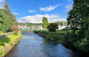 Den smukke flod Greta der løber gennem Keswick i Lake District med blå himmel, grønne træer og bjerge i baggrunden
