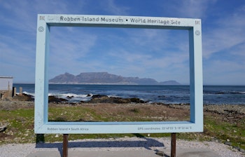 Træskilt med retningsanvisninger på Robben Island, Sydafrika, der peger mod forskellige seværdigheder, herunder det historiske fængselsområde, med Cape Town og Table Mountain i baggrunden