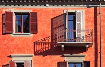 Traditionel rød husfacade med træskodder og balkon der kaster skygger i den historiske by Alba i Piemonte, Italien