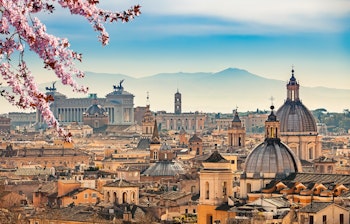 Betagende panoramaudsigt over Rom med blomstrende kirsebærtræer i forgrunden, historiske kupler og Vittoriano-monumentet badet i gyldent sollys, set fra Castel Sant'Angelo