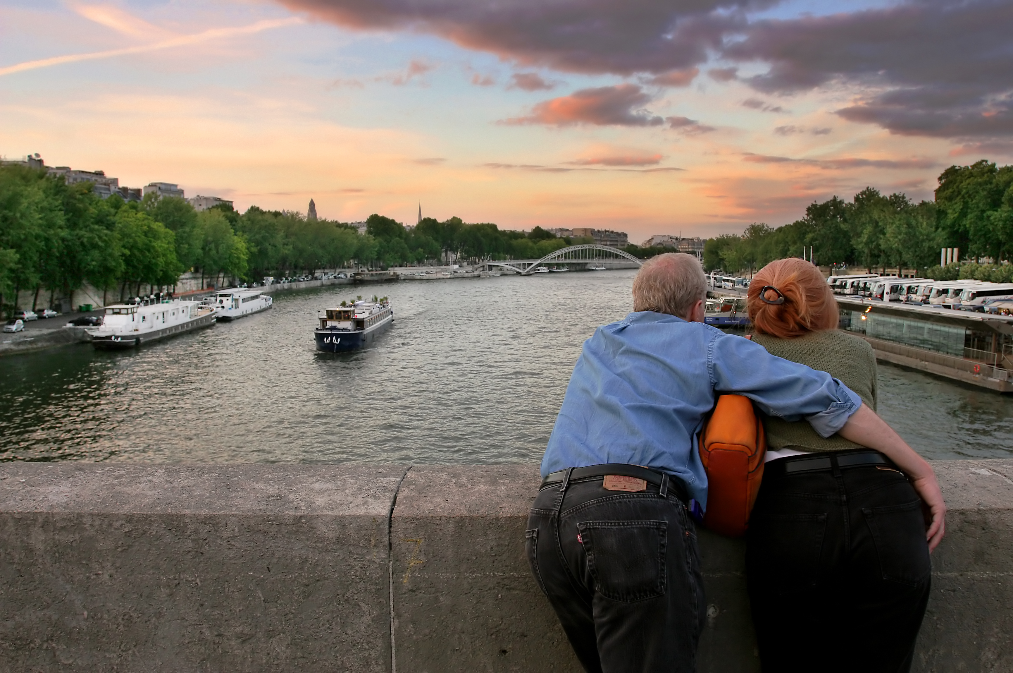 Ældre par nyder romantisk solnedgang over Seine-floden fra en bro i Paris med krydstogts-både