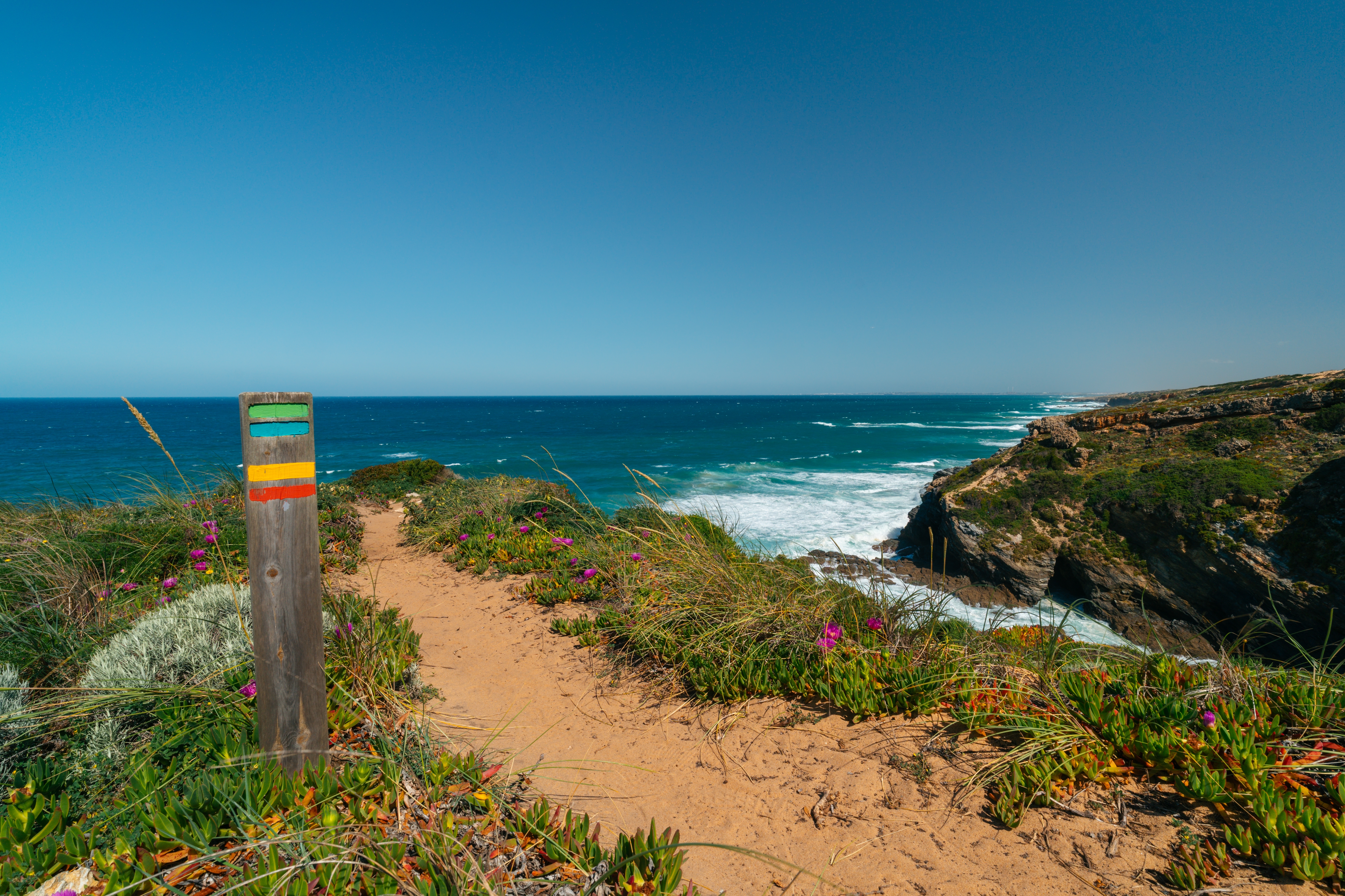 Farverig trailmarkering på Fishermans Trail langs Rota Vicentina i Portugal med dramatiske klippekyster og det blå Atlanterhav i baggrunden