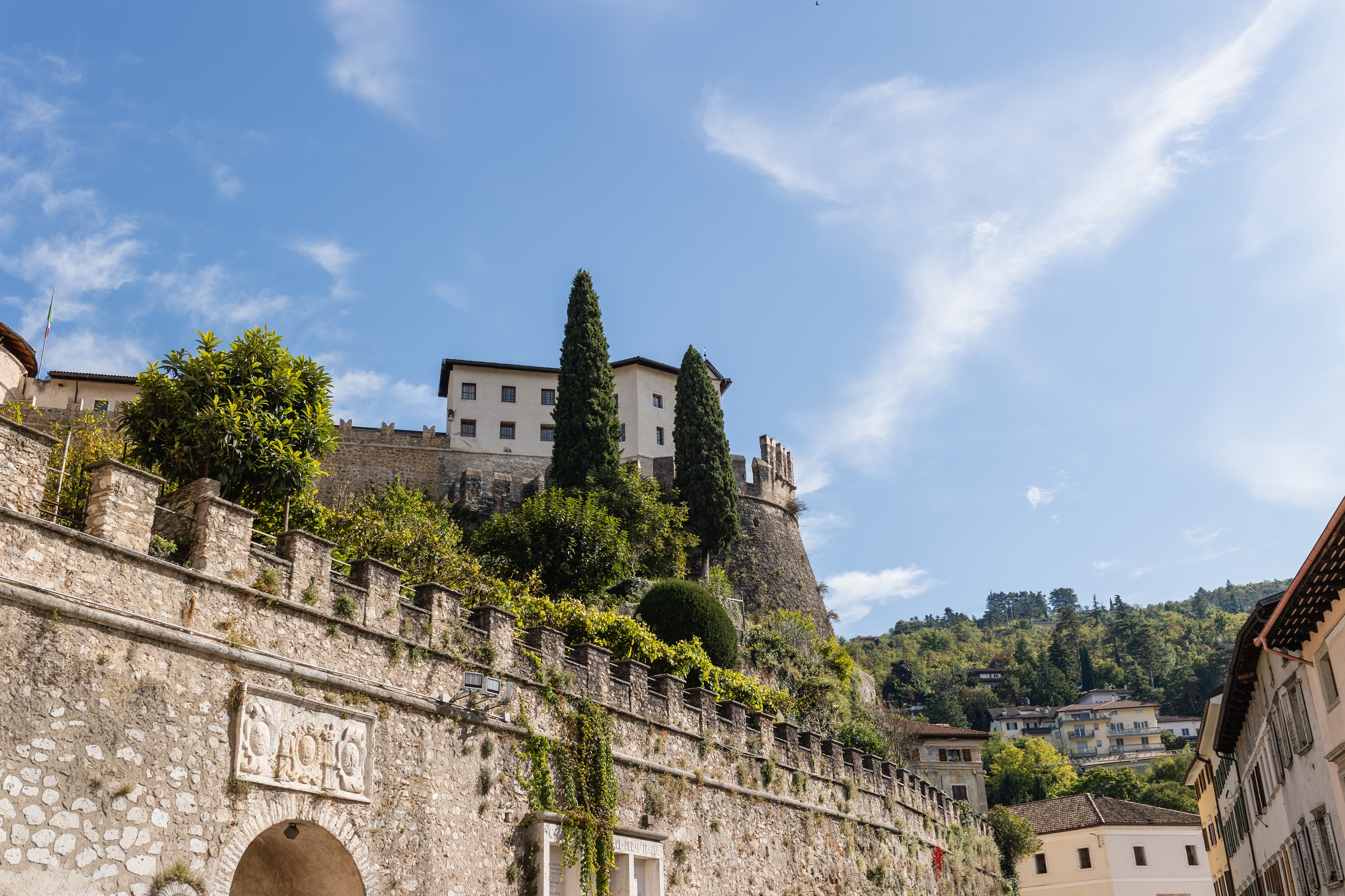 Udsigt nedefra af Rovereto Slot med historiske stenvolde og cyprestræer i Trentino, Italien, under en klar blå sommerhimmel