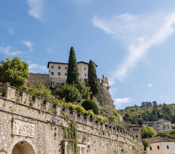 Udsigt nedefra af Rovereto Slot med historiske stenvolde og cyprestræer i Trentino, Italien, under en klar blå sommerhimmel