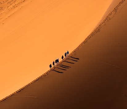 Luftfoto af rejsende der vandrer over gyldne sanddyner i Sahara ørkenen ved solnedgang