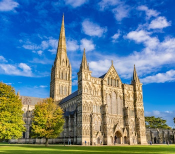 Den majestætiske Salisbury Katedral med sit høje gotiske spir omgivet af efterårstræer under en blå himmel i Wiltshire, England