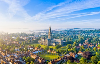 Betagende luftfoto af Salisbury Katedral der rejser sig gennem morgentågen med sin karakteristiske spir, omgivet af den historiske by i Wiltshire, England