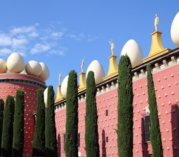 Det ikoniske Salvador Dalí Museum i Figueres med karakteristiske hvide æggeskulpturer på taget og cypresser foran den røde facade