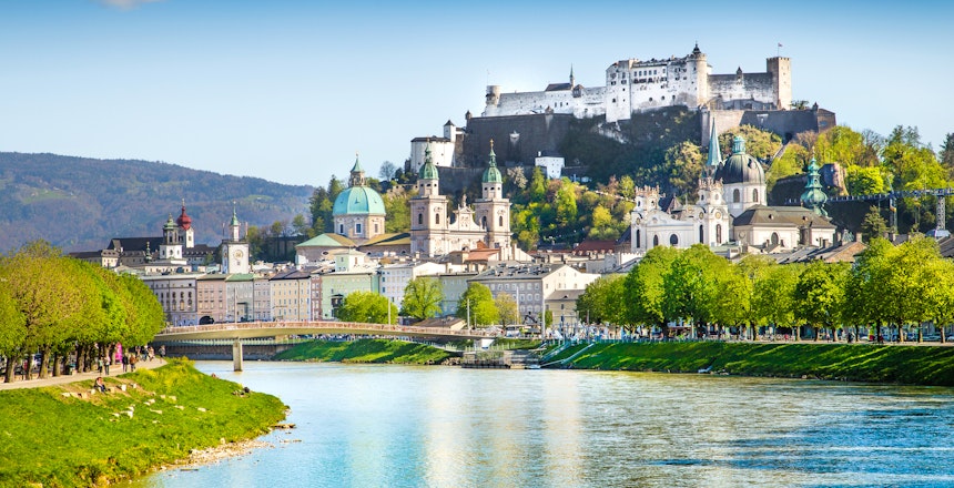 Malerisk panoramaudsigt over Salzburgs historiske bymidte med Hohensalzburg-fæstningen tronende på bjerget og Salzach-floden i forgrunden ved solnedgang