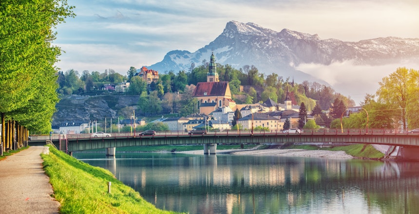 Betagende solnedgang over Salzburgs historiske bymidte med Hohensalzburg-fæstningen spejlet i Salzach-floden, badet i varmt gyldent lys med de østrigske alper i baggrunden