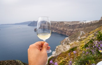 Hånd der holder et glas lokal Assyrtiko hvidvin med den betagende udsigt over Santorinis caldera, der viser øens vulkanske landskab og det blå Ægæerhav i Grækenland