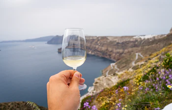 Hånd der holder et glas lokal Assyrtiko hvidvin med den betagende udsigt over Santorinis caldera, der viser øens vulkanske landskab og det blå Ægæerhav i Grækenland