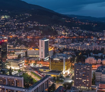 Panoramisk luftudsigt over Sarajevo ved skumring med oplyste bygninger, skyskrabere og omkringliggende bjerge i Bosnien-Hercegovina