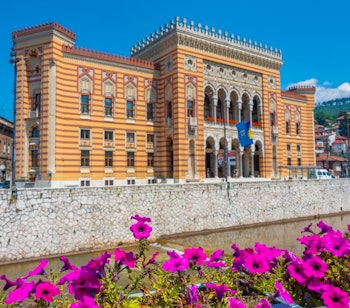 Det historiske Sarajevo Rådhus med sin imponerende facade ved Miljacka-floden omgivet af lyserøde blomster på en solrig dag i Bosnien-Herzegovina