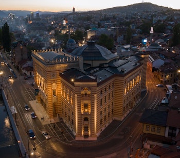 Luftfoto af det historiske Sarajevo Rådhus (Vijećnica) oplyst om natten med Miljacka-floden og byens panorama i Bosnien-Hercegovina