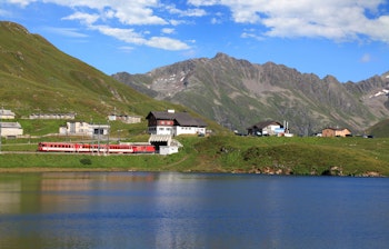 Rødt schweizisk bjergtog kører gennem alpelandskab med sø og snedækkede bjerge