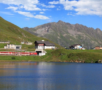 Rødt schweizisk bjergtog kører gennem alpelandskab med sø og snedækkede bjerge