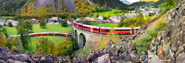 Rødt schweizisk tog kører over stenbro gennem alpine bjerge i Schweiz