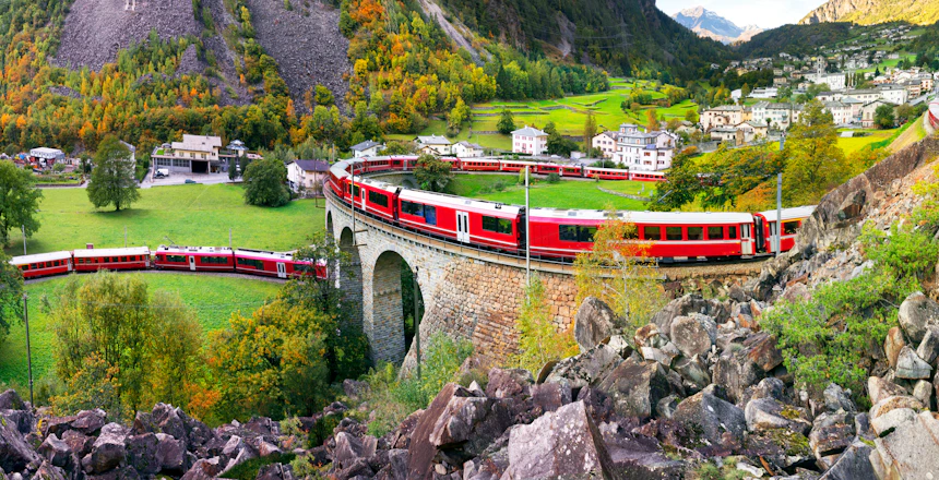 Rødt schweizisk tog kører over stenbro gennem alpine bjerge i Schweiz